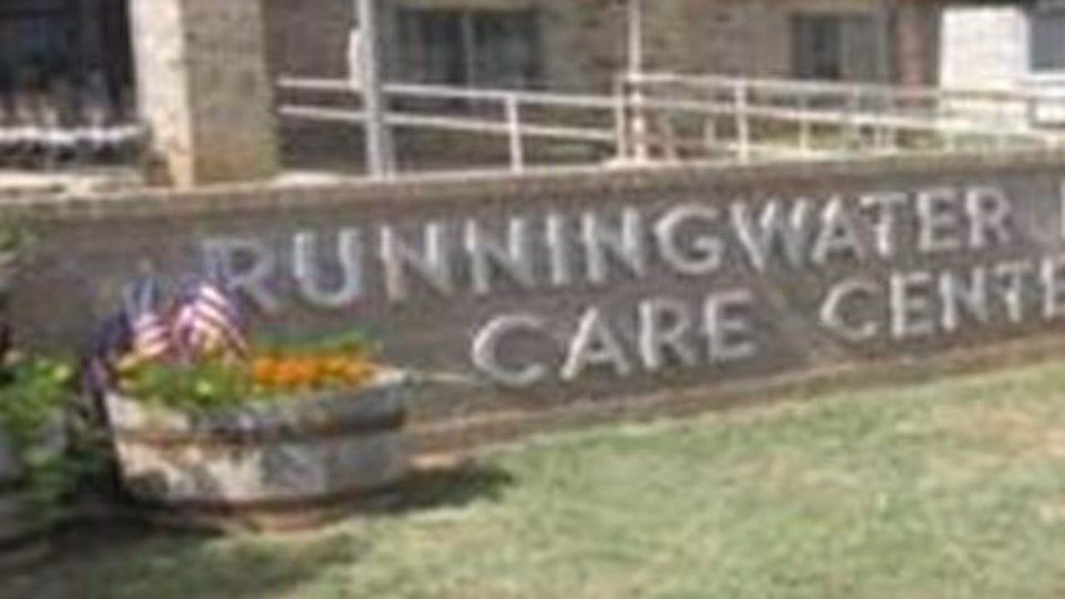 Stone sign with the text 'Runningwater Care Center' in front of a building, with a planter containing flowers and small American flags nearby.