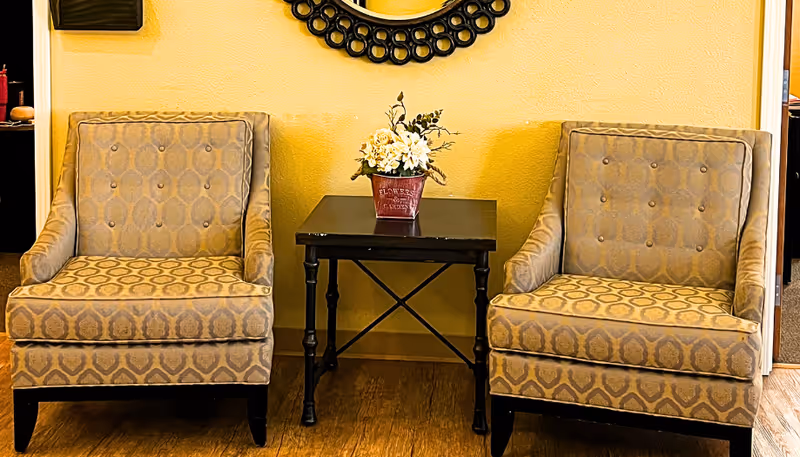 Two patterned armchairs with button tufting are positioned on either side of a small black side table holding a flower arrangement in a red pot. A decorative round mirror with a black frame hangs on the yellow wall behind the chairs.