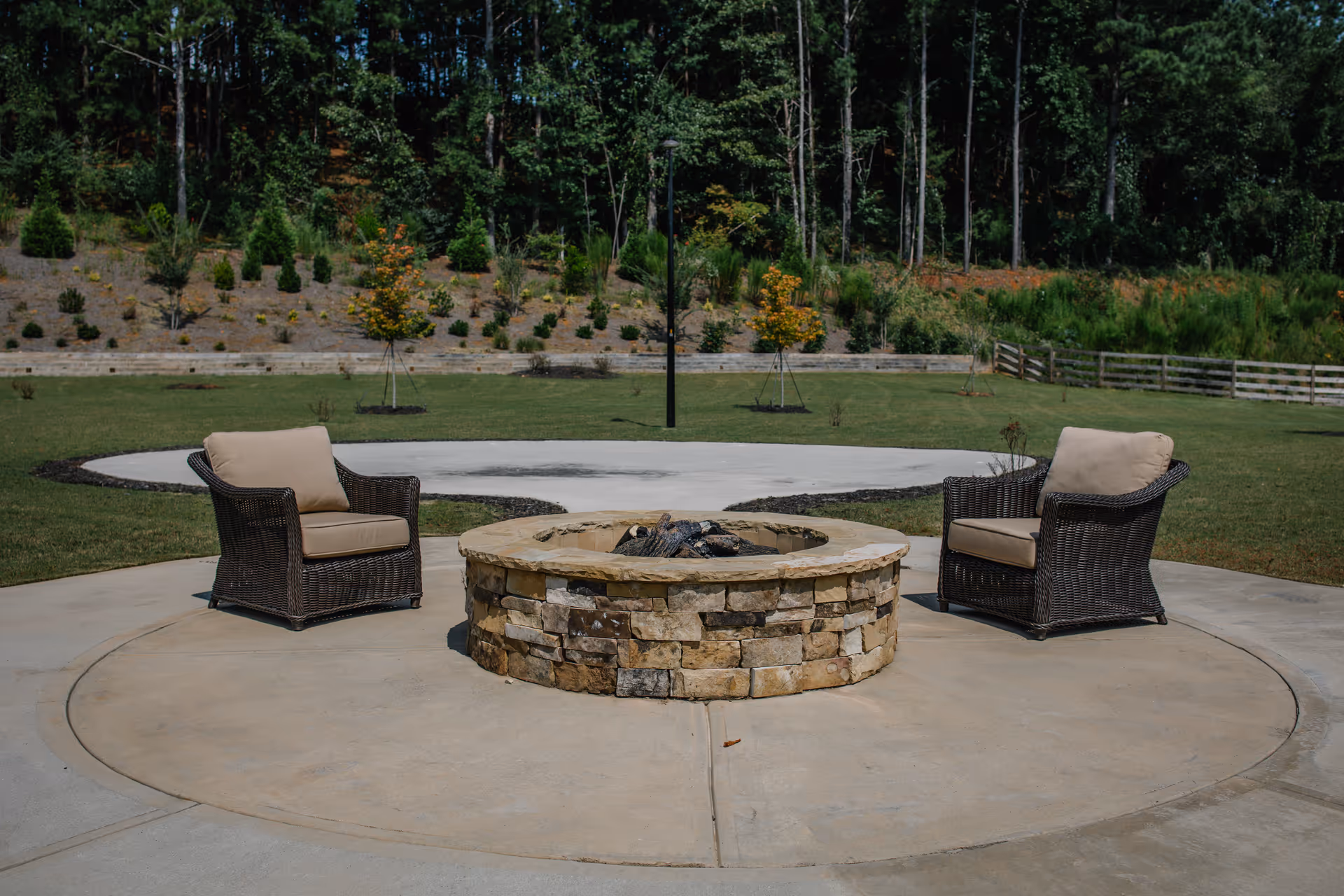 Outdoor seating area with two cushioned wicker chairs facing a circular stone fire pit, surrounded by a concrete patio. In the background, there is a grassy area with small trees and a wooded area beyond.