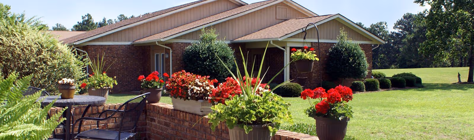 Outdoor patio area at Quail Haven Retirement Village featuring a brick wall with various potted plants and flowers, black metal chairs and a table, with a brick building and green lawn in the background under a clear sky.