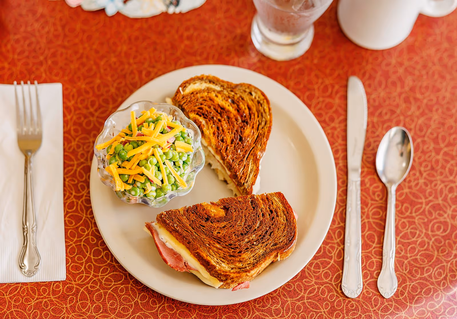 A plate with a grilled sandwich cut in half and a small glass bowl of pea salad topped with shredded cheddar cheese on a red patterned tablecloth. Silverware including a fork on a napkin, a knife, and a spoon are placed around the plate.