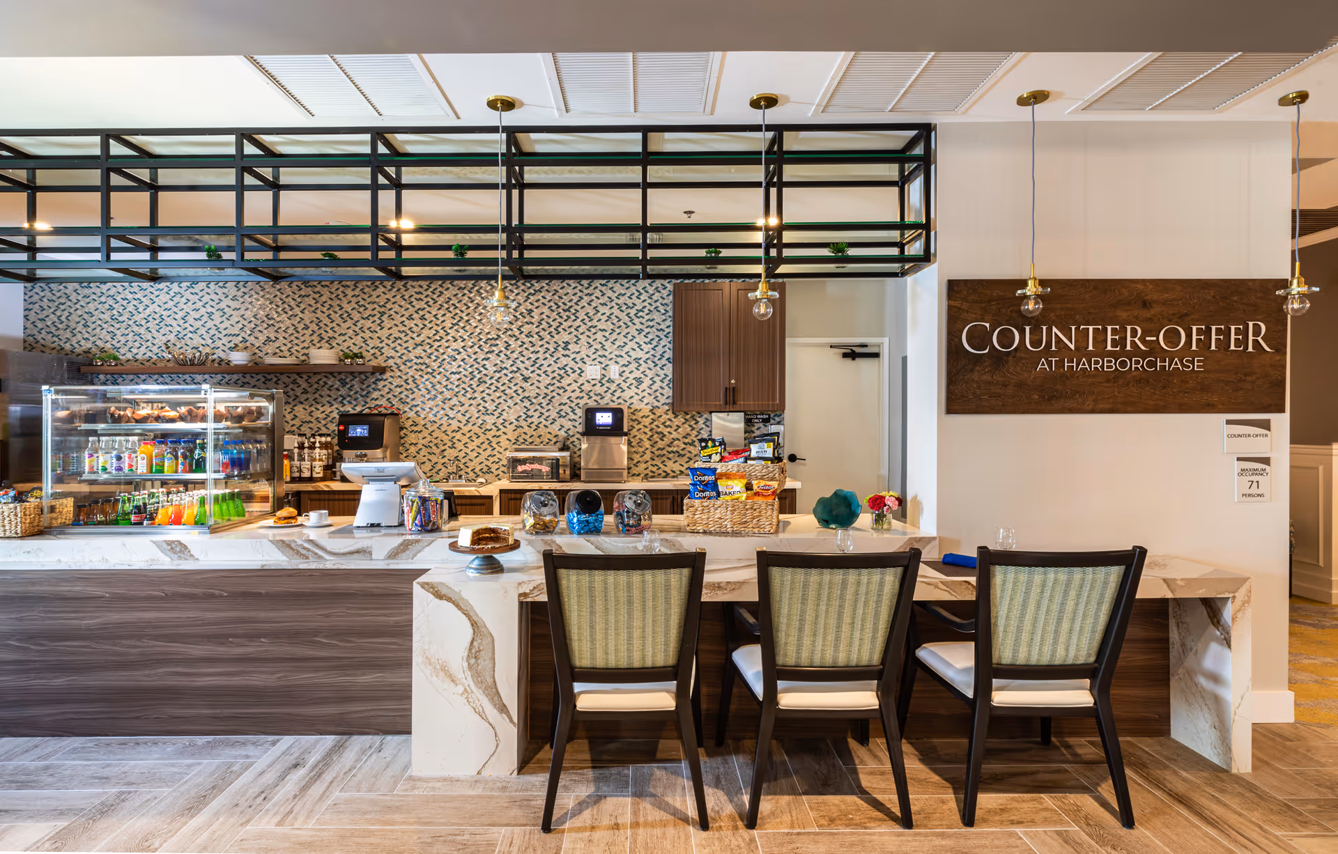 A modern snack and beverage counter area with three chairs in front. The counter has a marble surface with snacks, drinks, and a cake on display. Behind the counter is a patterned tile wall with shelves, a coffee machine, and cabinets. A wooden sign on the wall reads 'COUNTER-OFFER AT HARBORCHASE'.