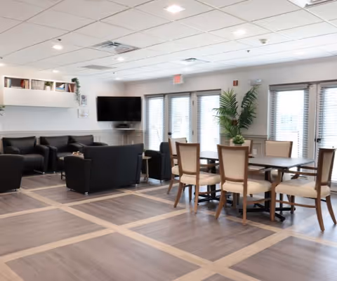 A spacious and well-lit common area featuring a seating arrangement with black armchairs and sofas on the left, a flat-screen TV mounted on the wall, and a dining table with six chairs on the right. Large windows with blinds allow natural light to fill the room, and a tall green plant adds a touch of nature near the dining area.