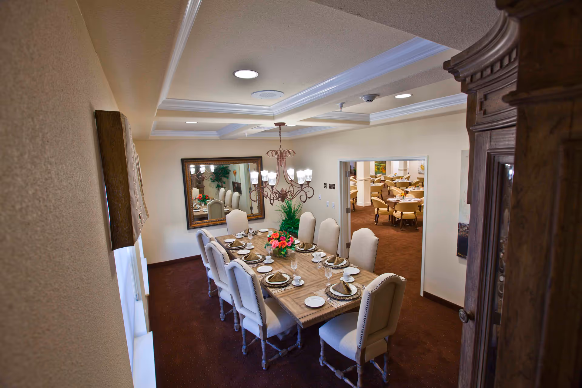 A formal dining room with a long set table, upholstered chairs, a chandelier, and a mirror on the wall with a view into a larger dining area.