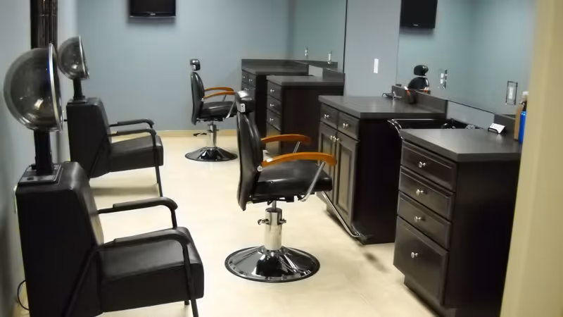 Interior view of a salon area with black salon chairs, hair dryer stations, and dark wood cabinetry with mirrors on the wall.