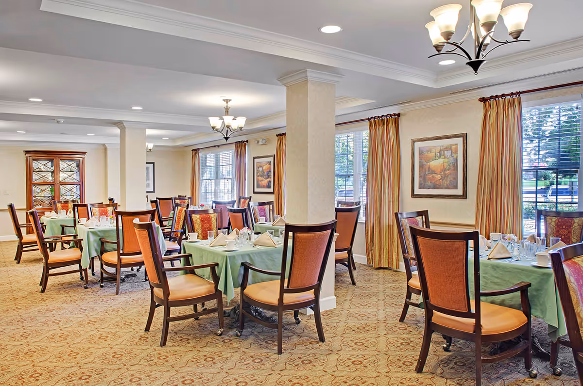 A bright and elegant dining room with multiple tables covered in green tablecloths, each set with napkins, glasses, and cups. The room features wooden chairs with orange cushions, large windows with striped curtains, framed artwork on the walls, and ceiling light fixtures.