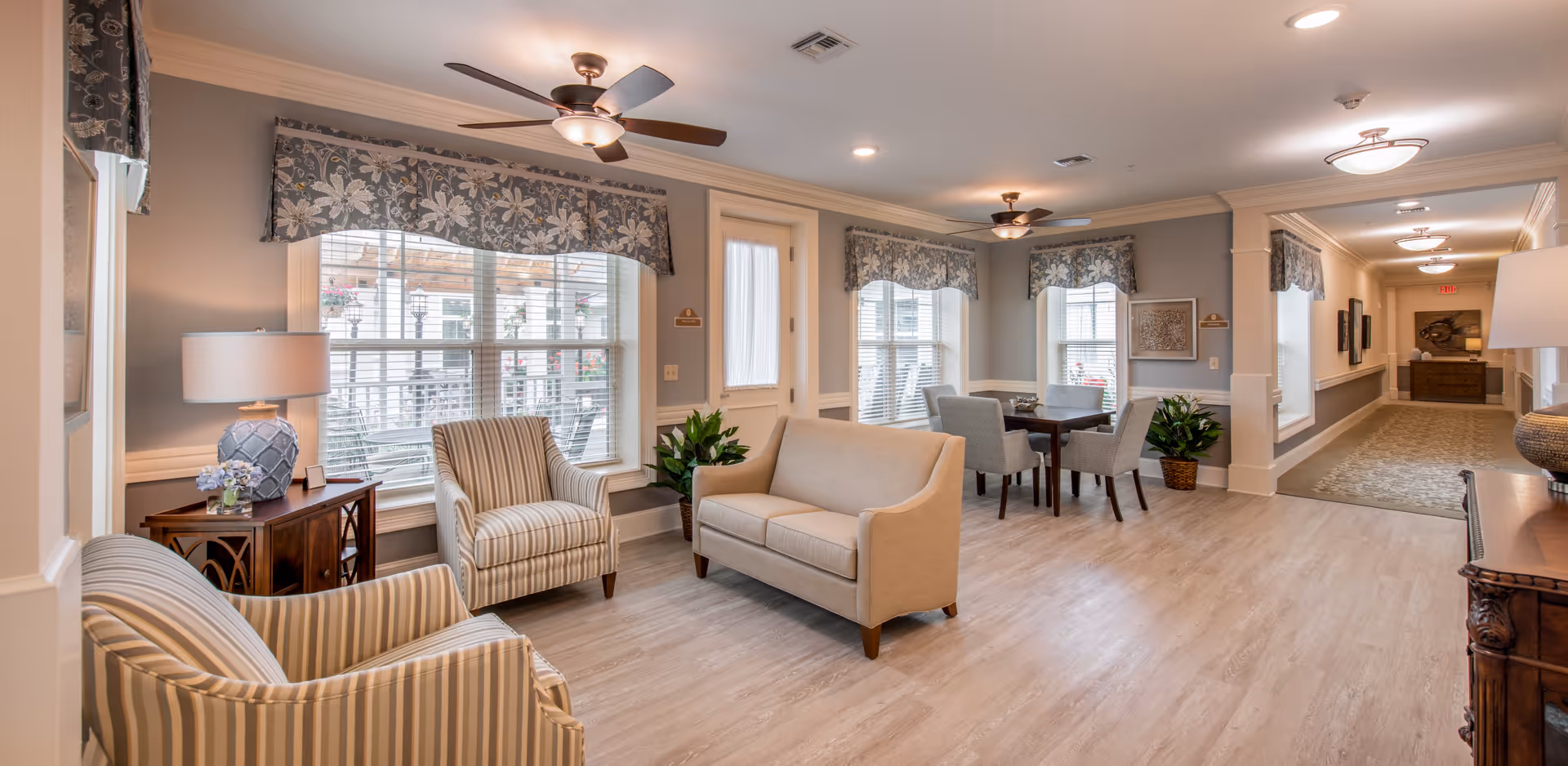 A bright and spacious common area in a senior living facility featuring a beige loveseat, two striped armchairs, a wooden side table with a lamp and flowers, and a dining table with four chairs near large windows with floral valances. The room has light wood flooring, soft gray walls, ceiling fans with lights, and a hallway leading further into the facility.