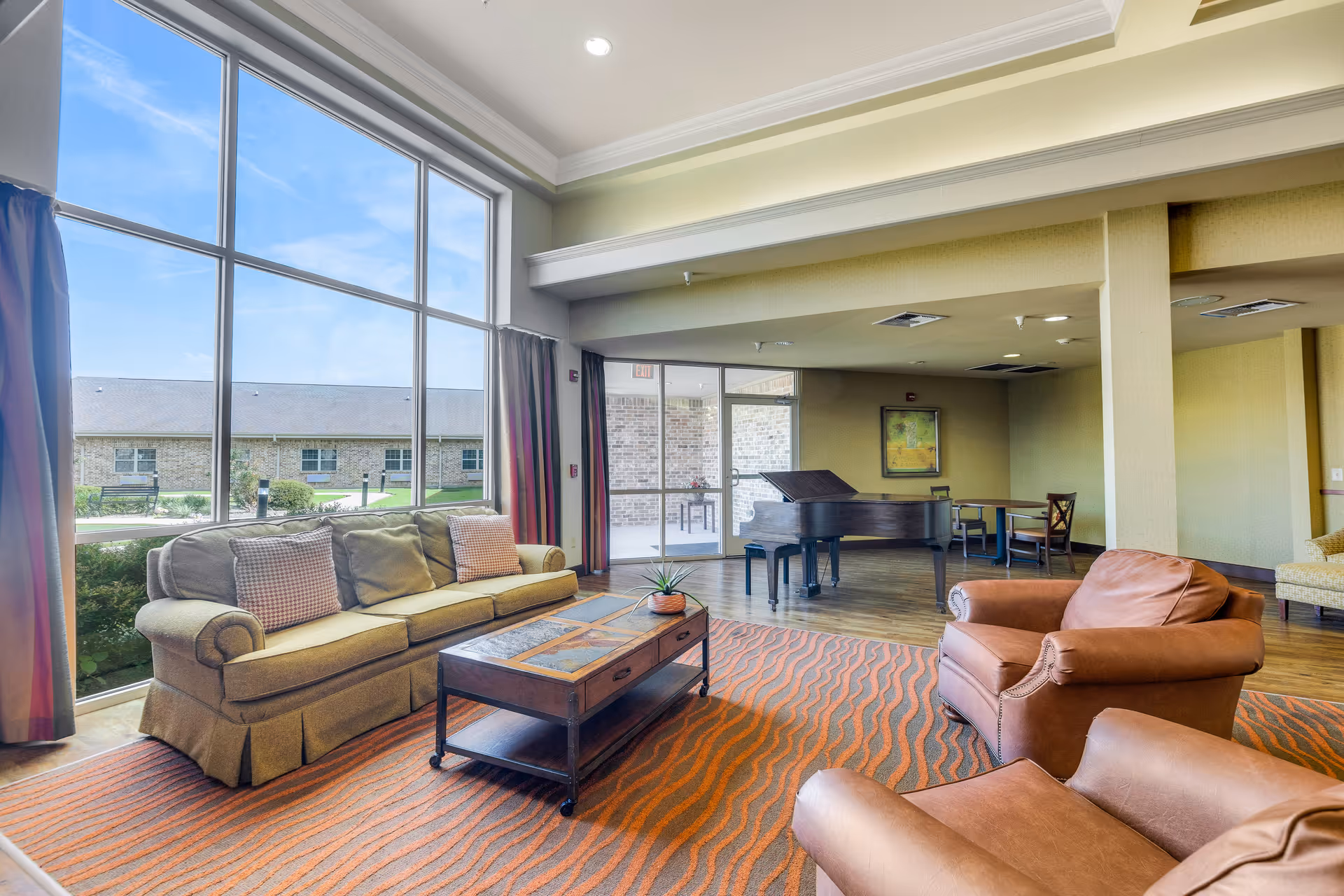 Sunlit common lounge with a sofa, leather armchairs, coffee table and a grand piano beside large floor-to-ceiling windows.