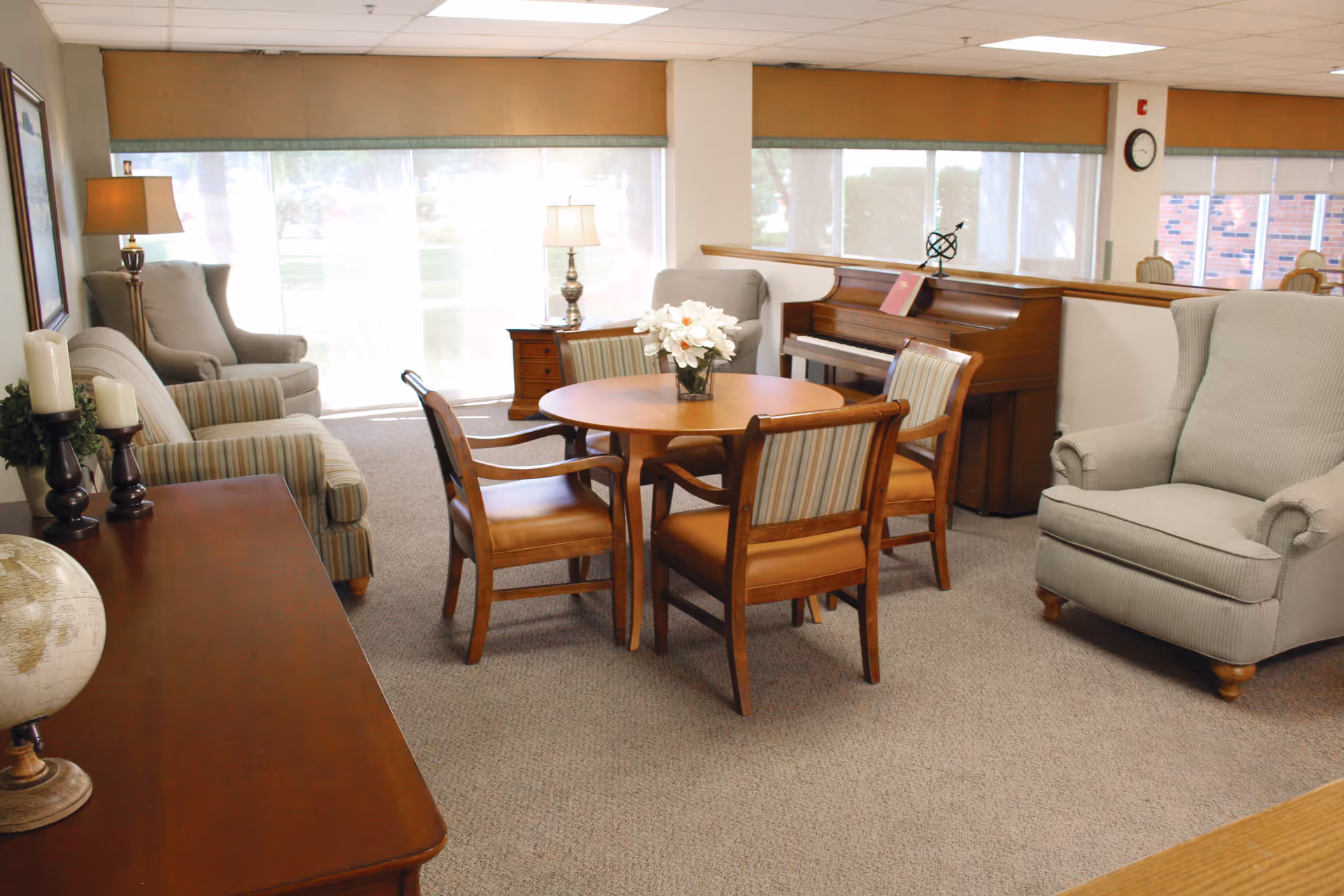 A bright and cozy living room area in a senior living facility with large windows covered by blinds. The room features a round wooden table with four chairs, a piano, two armchairs, a sofa, a side table with a lamp, and a wooden console table with decorative candles and a globe.