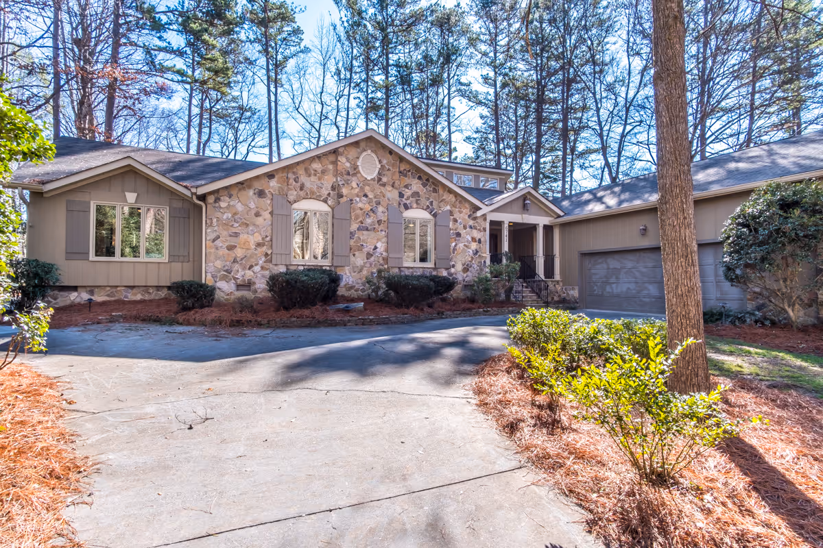 Exterior view of a single-story residential building with a stone facade and beige siding, surrounded by trees and shrubs. The driveway leads to a two-car garage on the right side of the house. The scene is set on a clear, sunny day.