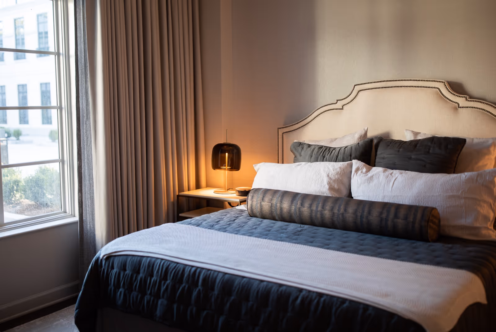 A cozy bedroom with a neatly made bed featuring multiple pillows and a cylindrical bolster pillow. The bed has a beige upholstered headboard with nailhead trim. Next to the bed is a small nightstand with a modern dark-colored lamp emitting warm light. A large window with beige curtains allows natural light to enter the room.