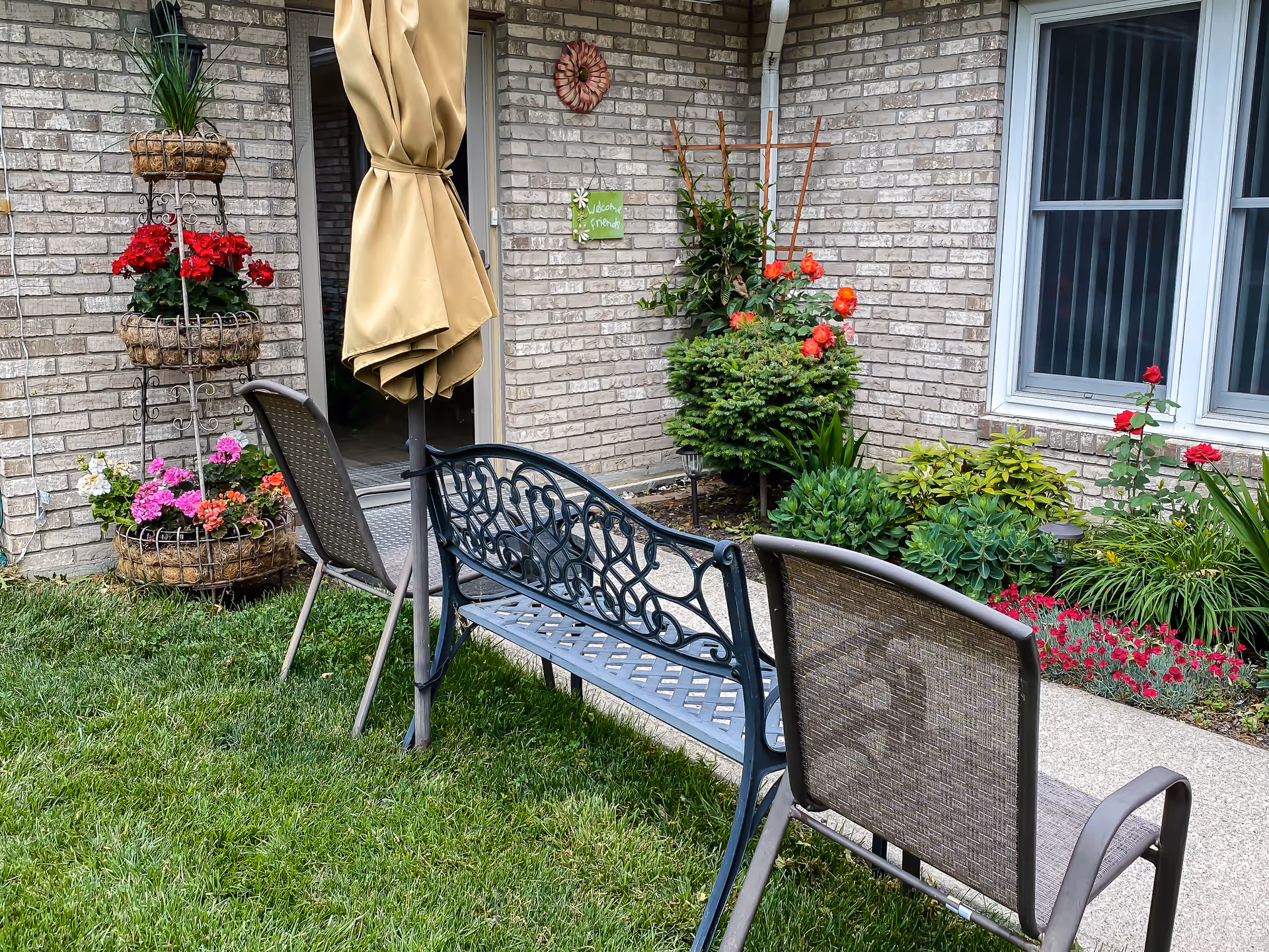Outdoor patio area with a metal bench and two chairs on a grassy lawn. There is a closed beige umbrella attached to the bench. The background shows a brick wall with a door, windows, and various colorful flowers and plants in garden beds and hanging baskets.