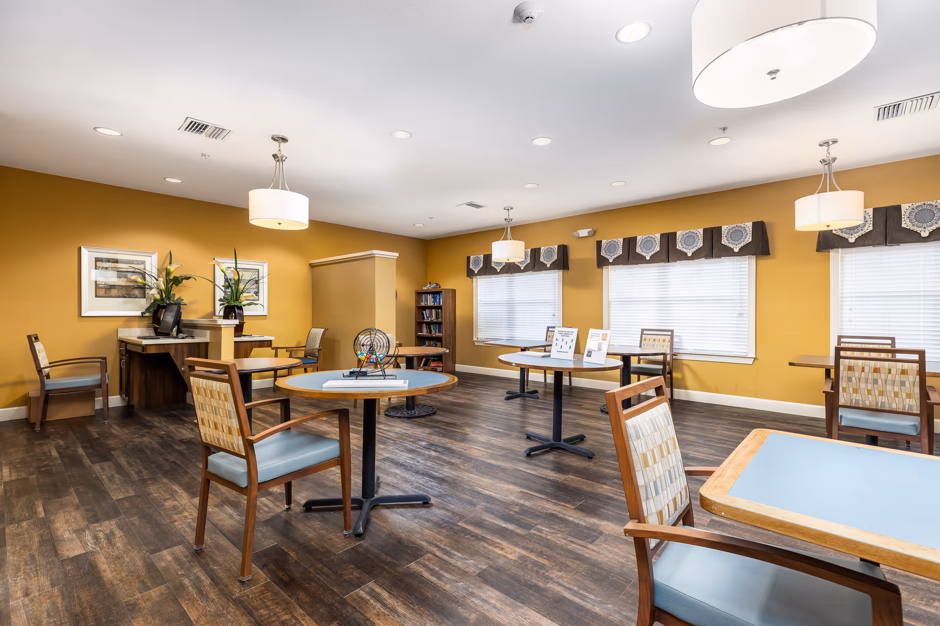 A spacious room with several round and square tables surrounded by wooden chairs with cushioned seats. The walls are painted mustard yellow, and there are three large windows with white blinds and decorative valances. The room has wooden flooring and is well-lit with ceiling lights and hanging lamps. There are framed pictures and plants on a counter along one wall, and a bookshelf in the corner.