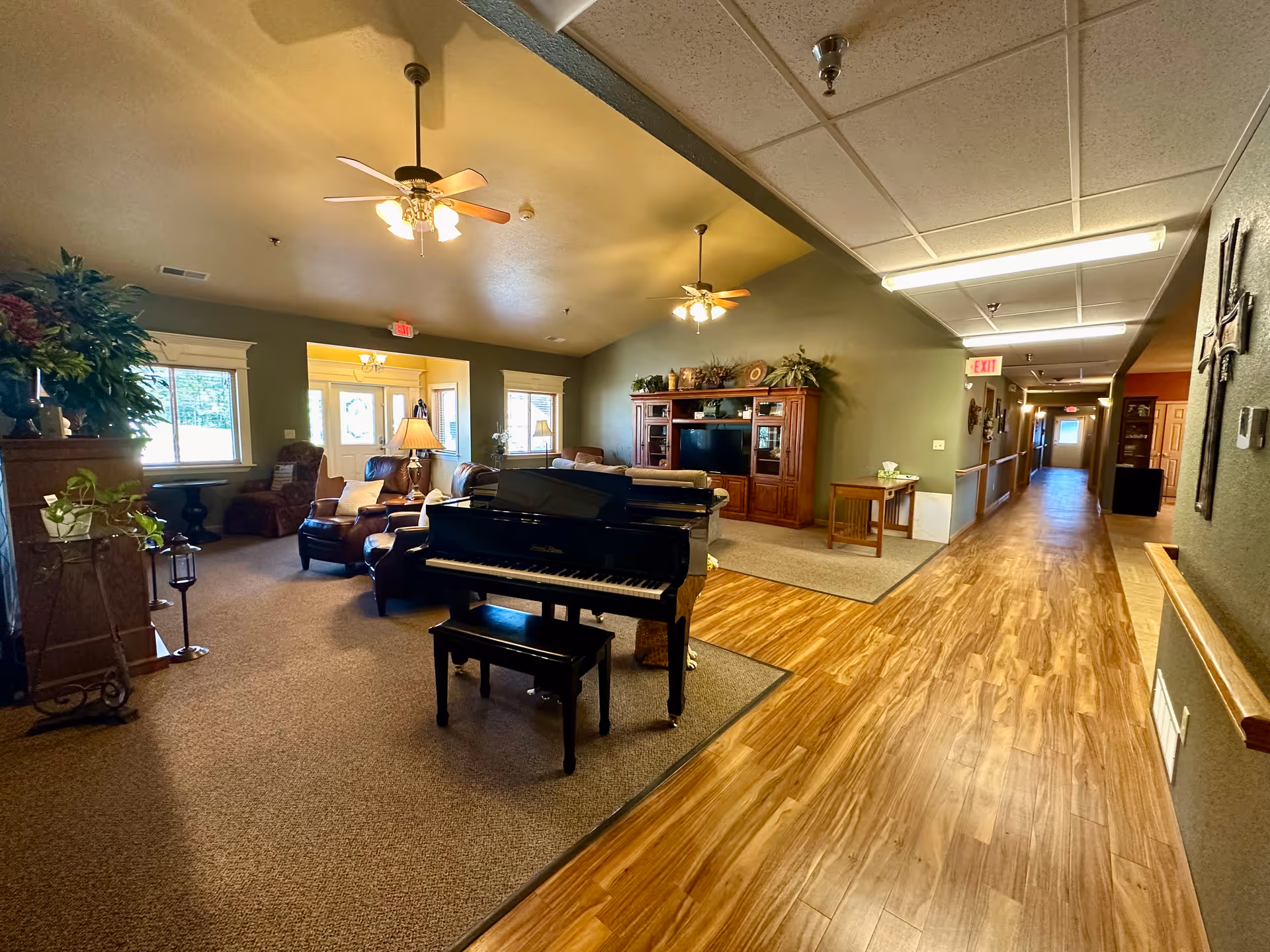 Interior view of a senior living facility common area with a black grand piano in the foreground, comfortable seating including leather chairs and sofas, a wooden entertainment center with a TV, ceiling fans with lights, and a long hallway with wooden flooring extending to the right.