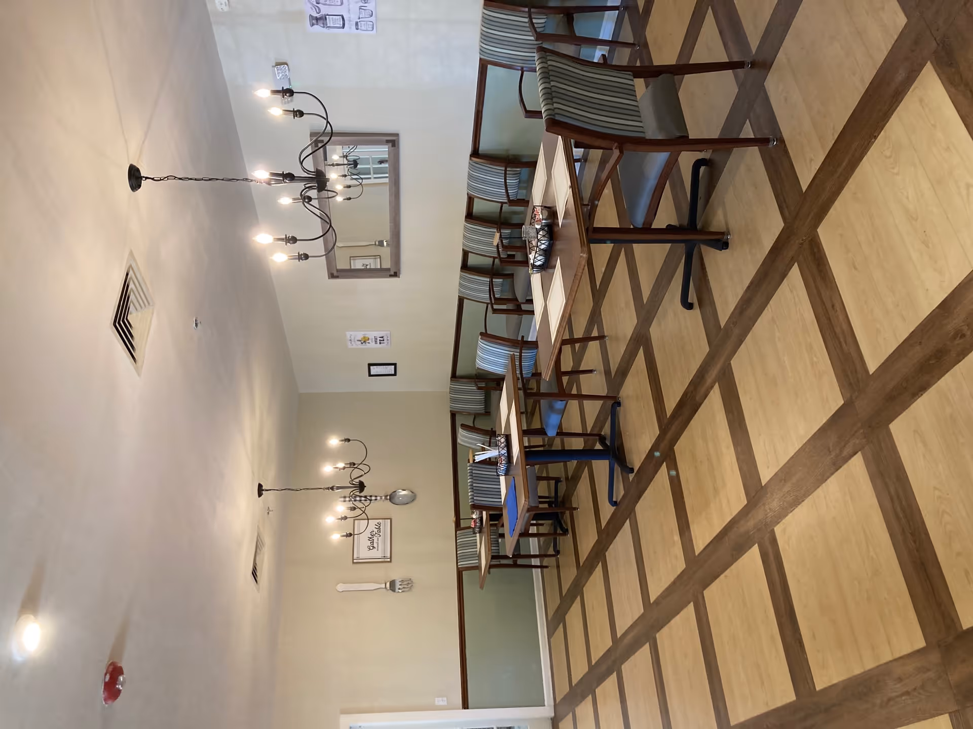 A dining room with wooden tables and chairs arranged neatly. The room has a checkered wood and light brown floor, light green and beige walls, and two black chandeliers with lit bulbs hanging from the ceiling. There are framed pictures and decorative oversized utensils on the walls, and a large mirror reflecting part of the room.