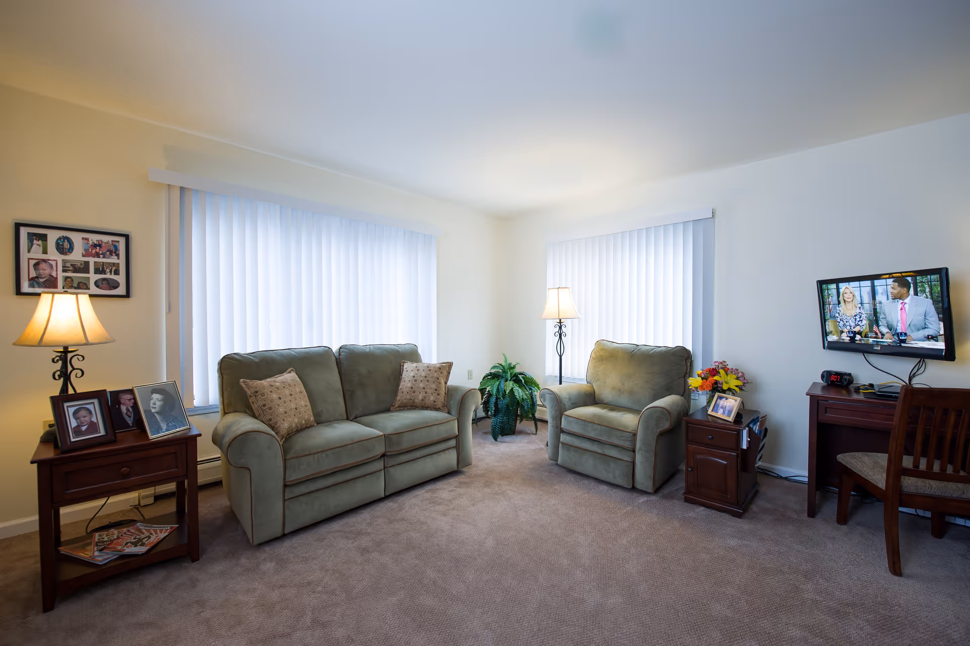 Bright living room with matching green sofa and armchair, side tables with lamps and framed photos, a TV on a desk, and large vertical blinds on the windows.