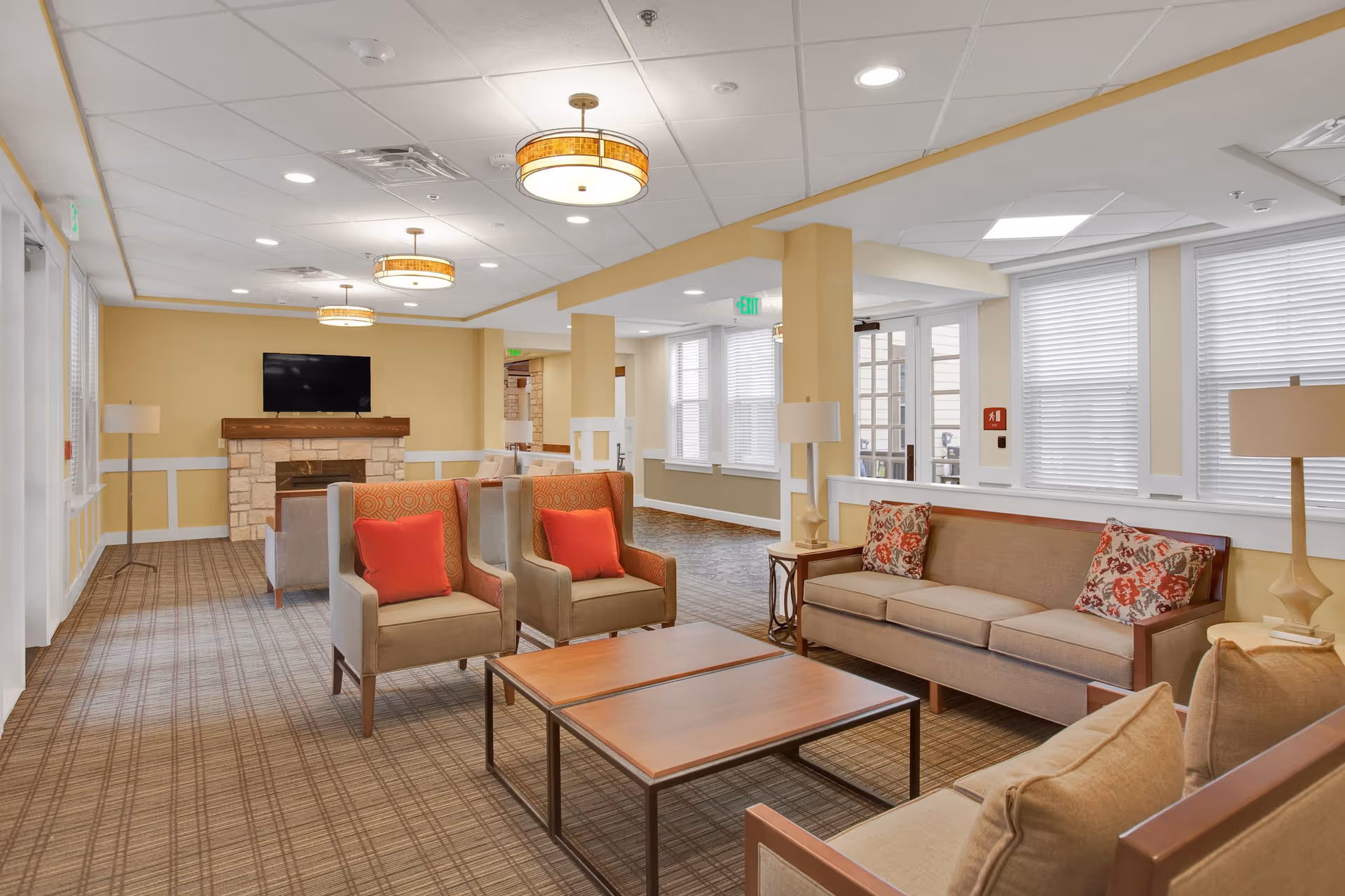 Well-lit communal living room with sofas, armchairs, coffee tables, a TV over a fireplace, and large windows.