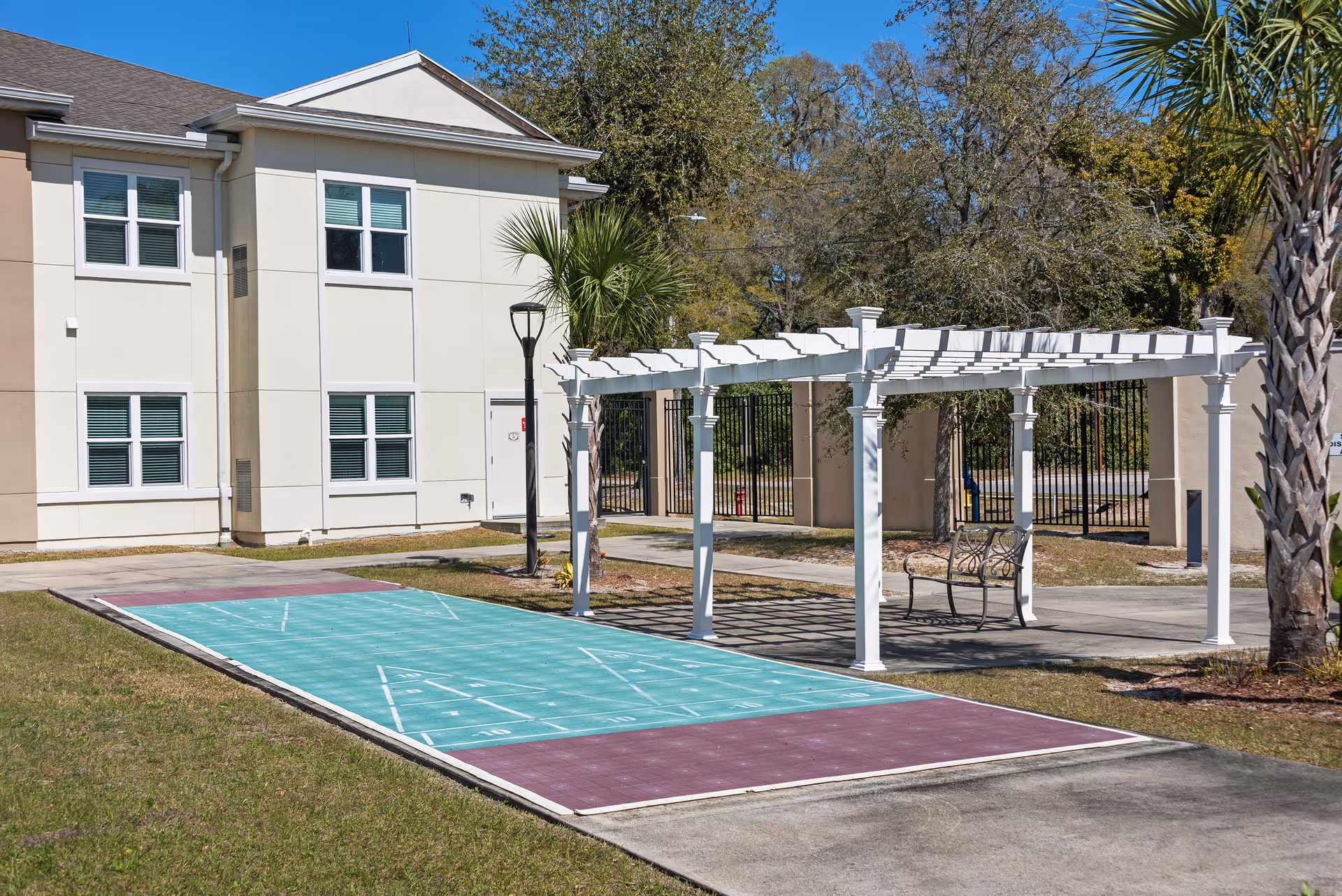 Outdoor area at Blue Palms Senior Living of Deland featuring a shuffleboard court with a white pergola and a bench nearby. The background shows part of the senior living building, palm trees, and a clear blue sky.