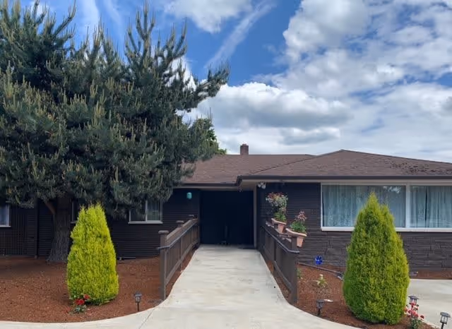 Exterior view of a single-story building with a brown roof and dark brown walls. A concrete ramp with wooden railings leads to a covered entrance. There are two tall green shrubs on either side of the ramp and a large pine tree to the left. The sky is partly cloudy with patches of blue.