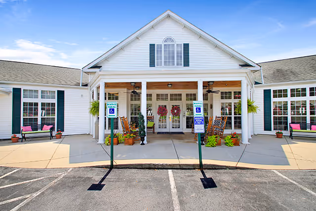 Front entrance of a white senior living facility with a covered porch, rocking chairs, potted plants, and accessible parking signs.