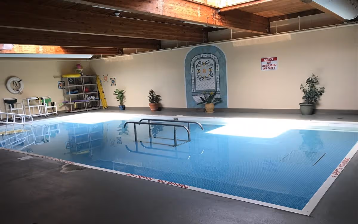 Indoor swimming pool with clear blue water, metal handrails, potted plants along the wall, a decorative wall mural, and a sign that reads 'NOTICE NO LIFEGUARD ON DUTY'. The ceiling has exposed wooden beams and skylights allowing natural light to enter.