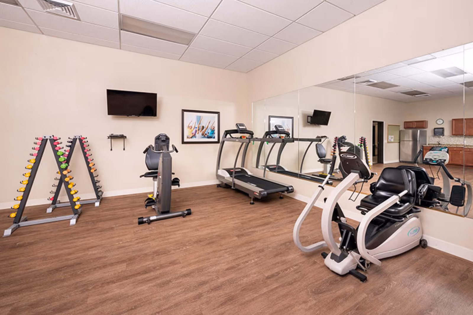 A fitness room with exercise equipment including a recumbent bike, treadmill, and a rack of colorful dumbbells. The room has wood flooring, a large wall mirror, a wall-mounted TV, and a framed picture on the wall. In the background, there is a kitchenette area with cabinets and a refrigerator.