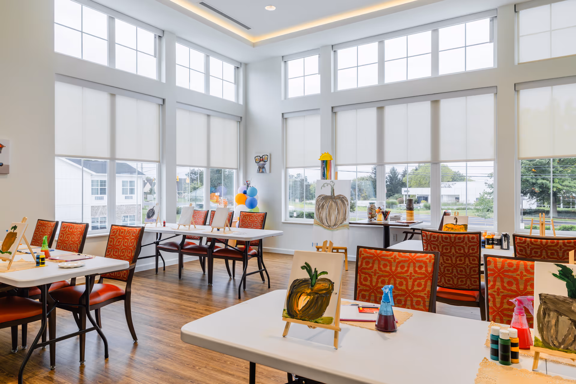 Bright and spacious activity room with large windows and white roller shades, featuring tables with red patterned chairs arranged for painting activities. Several small easels display paintings of pumpkins, and various art supplies are placed on the tables. The room has wooden flooring and a high ceiling with recessed lighting.