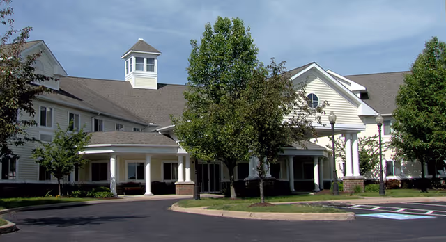 Exterior front view of Canterbury Senior Care Facility showing a large, light-colored building with multiple windows, a covered entrance supported by white columns, a small tower structure on the roof, and several trees and landscaped areas in front. The sky is clear with some clouds.