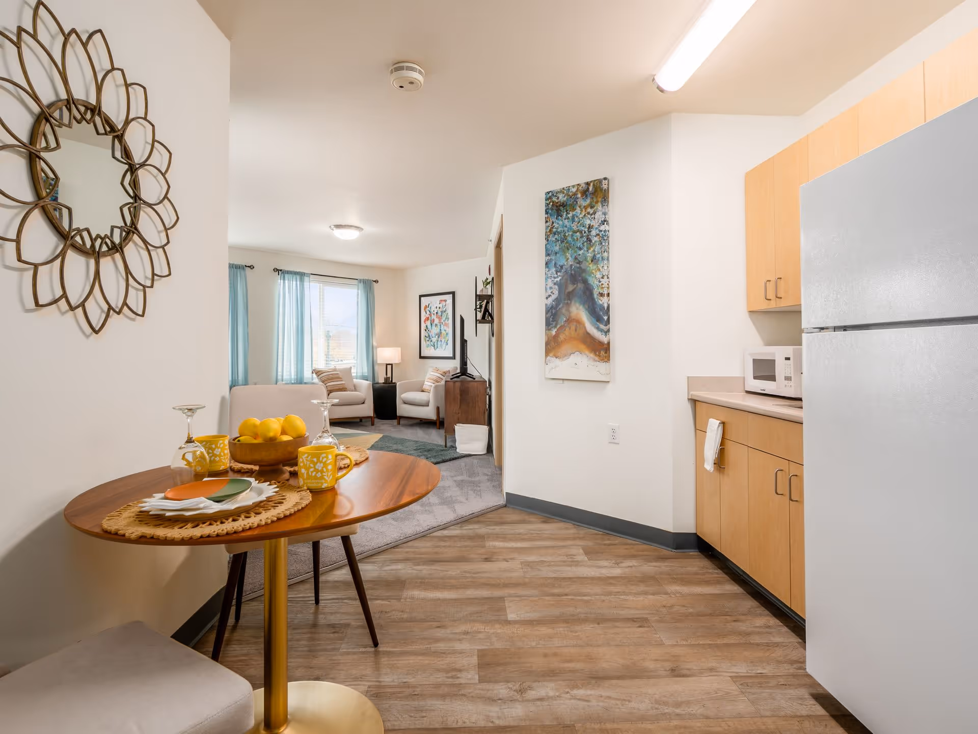 A cozy interior space featuring a small round wooden dining table set with plates, mugs, and a bowl of lemons. To the right is a kitchenette with light wood cabinets, a microwave, and a refrigerator. In the background, a living area with two armchairs, a TV on a stand, and windows with light blue curtains is visible. The walls are white, and there is a decorative mirror on the left wall and a colorful abstract painting on the right wall.