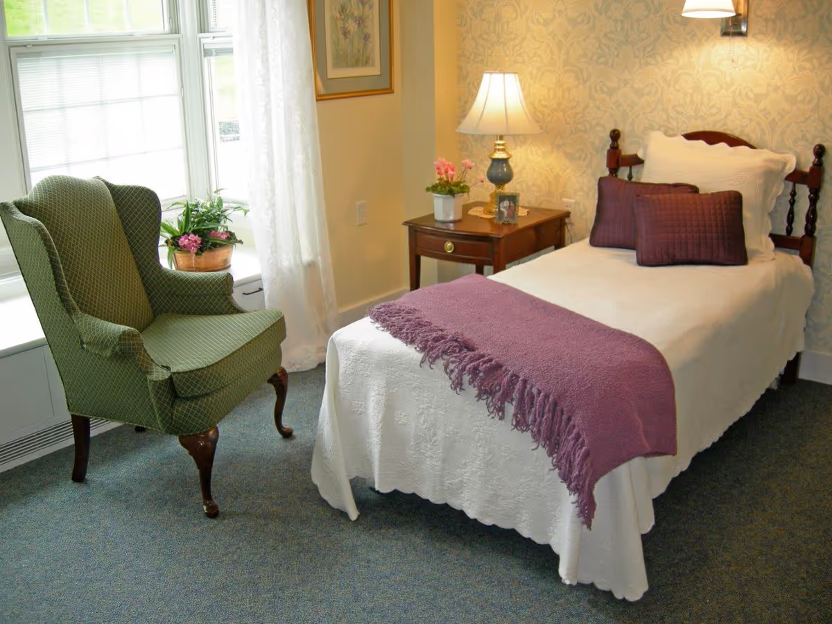 A cozy bedroom with a single bed covered in white bedding and a purple throw blanket. There are two purple pillows and one white pillow on the bed. Next to the bed is a wooden nightstand with a lamp, a small potted plant, and a framed photo. A green upholstered armchair is positioned near a window with sheer white curtains, and a potted plant sits on the windowsill. The walls have a light patterned wallpaper.