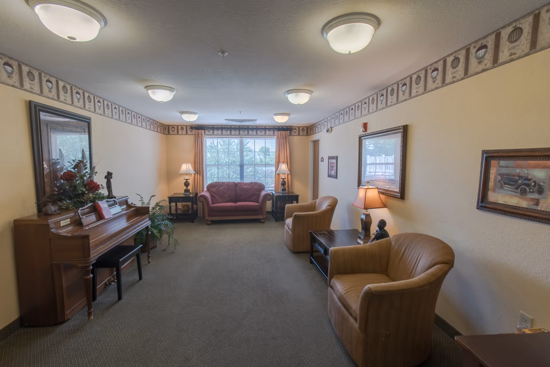 A cozy living room with a piano on the left side adorned with a flower arrangement, a red loveseat in front of a large window with curtains, two beige armchairs, side tables with lamps, framed artwork on the walls, and a carpeted floor.