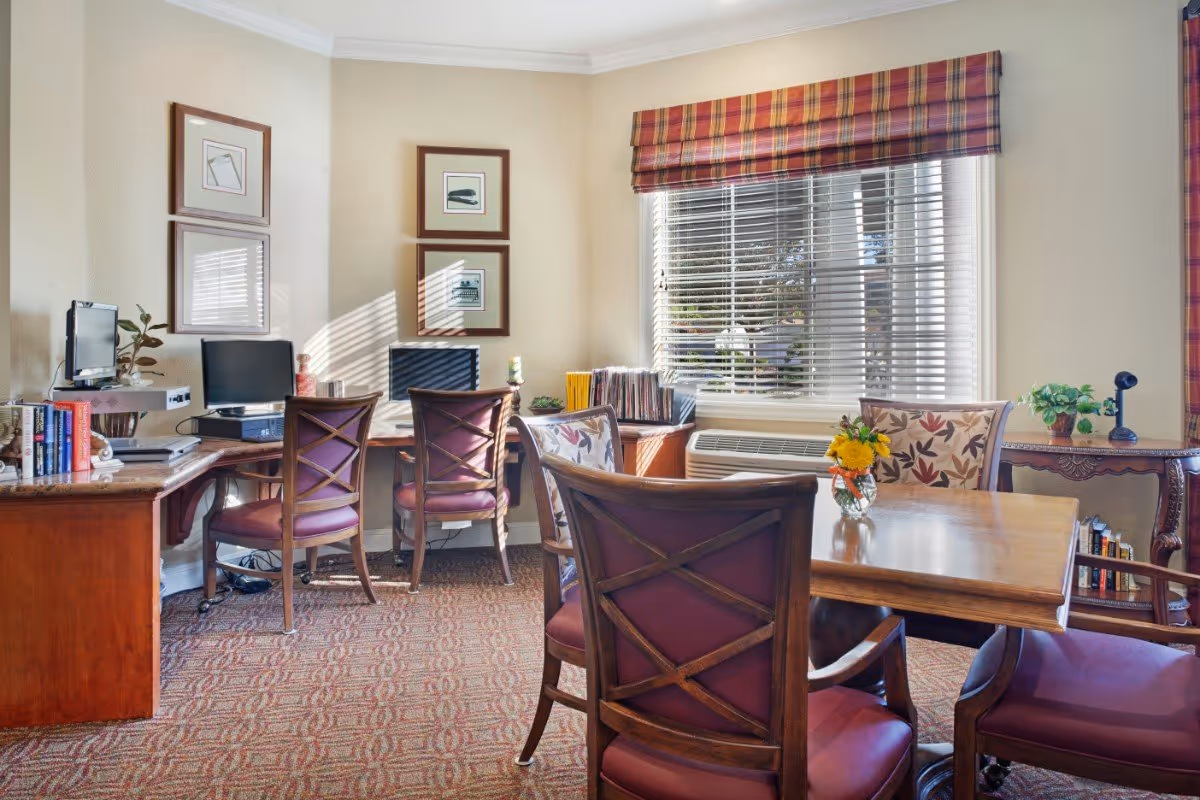 A cozy interior room with a wooden table surrounded by chairs, a window with blinds and a plaid valance, two computer desks with chairs and desktop computers, framed artwork on the walls, and decorative plants and books on the furniture.