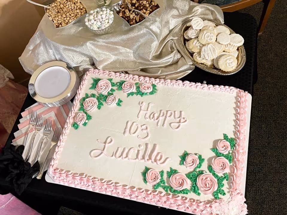 A rectangular birthday cake decorated with pink roses and green leaves, with the message 'Happy 103 Lucille' written in pink icing. The cake is placed on a black tablecloth with a silver cloth behind it. Nearby are plates, forks, napkins, bowls of nuts, and a silver tray with white cookies.