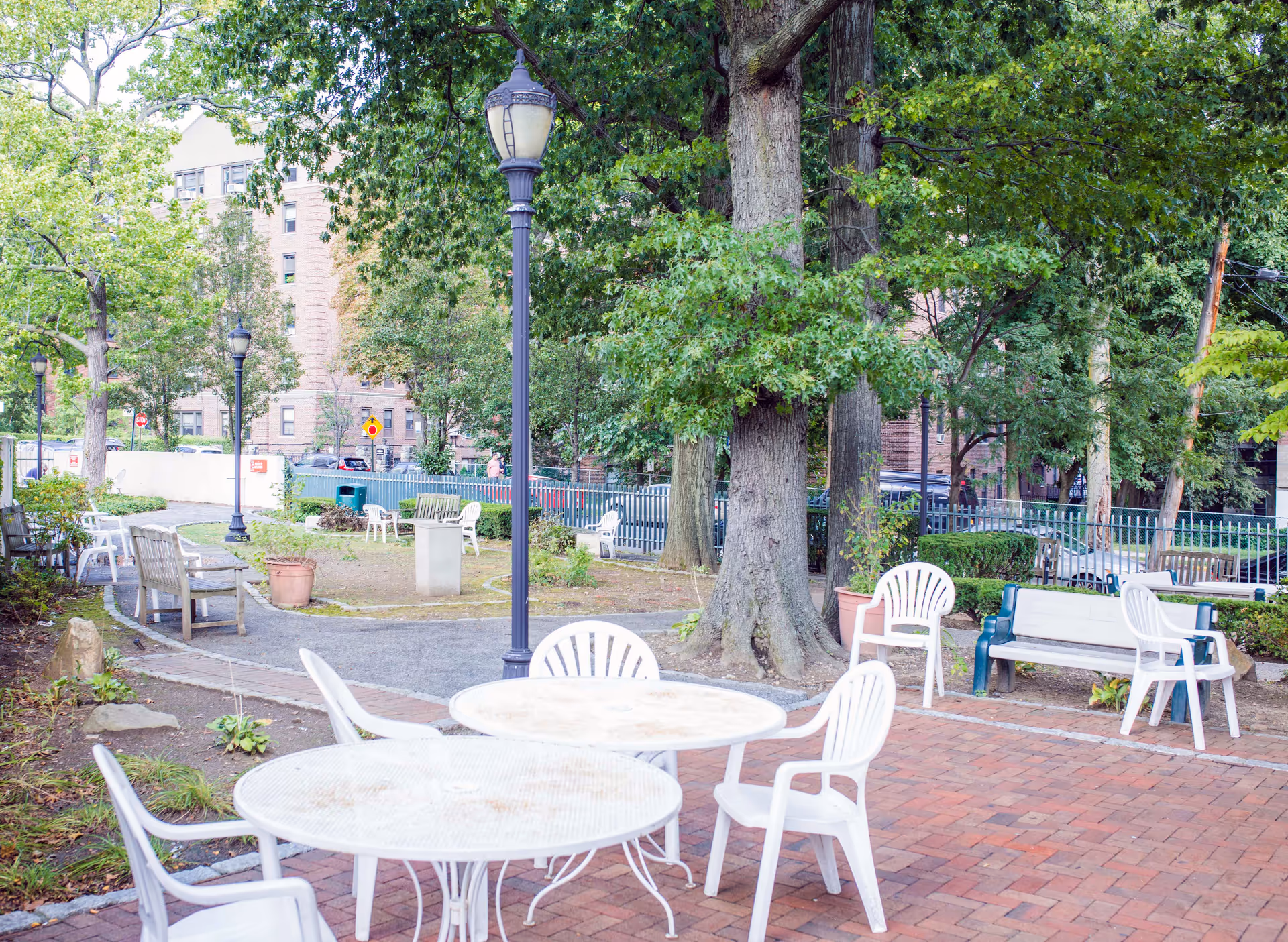 Outdoor courtyard patio with round tables, white plastic chairs, benches, large trees and lamp posts near a multi-story building.