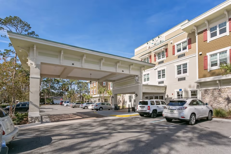 Exterior view of Beach House Assisted Living & Memory Care building with a covered entrance and several parked cars in the parking lot under a clear blue sky.