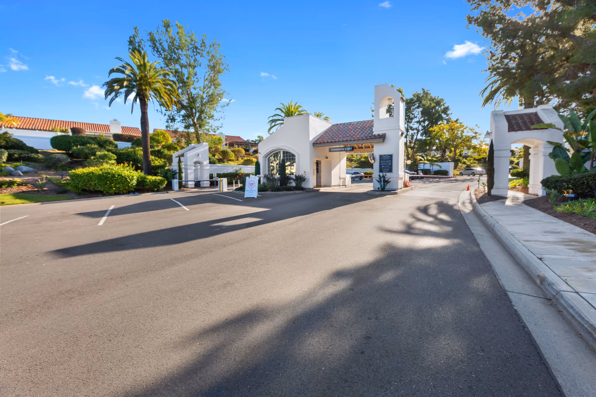 Entrance gate of Ocean Hills Country Club with white stucco buildings and red tile roofs, surrounded by palm trees and landscaped greenery under a clear blue sky.