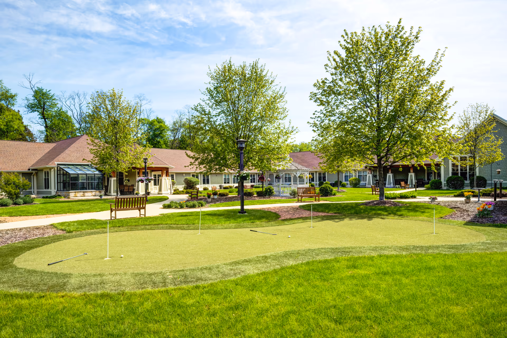 Sunlit courtyard with a putting green, benches, trees, and single-story senior living buildings in the background.