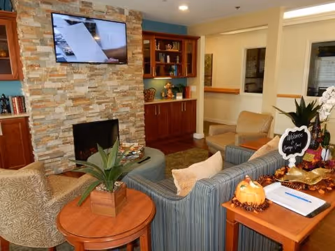 A cozy living room area with a stone fireplace and a mounted flat-screen TV above it. The room features a striped sofa, two armchairs, a round wooden side table with a potted plant, and a wooden cabinet with shelves and decorative items. A small table with a pumpkin decoration, a welcome sign, and a clipboard is also visible.