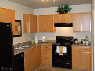 A kitchen with light wood cabinets, a black refrigerator, black stove with a white and orange towel hanging on the handle, a double sink, and various decorative items on the countertops and above the cabinets.