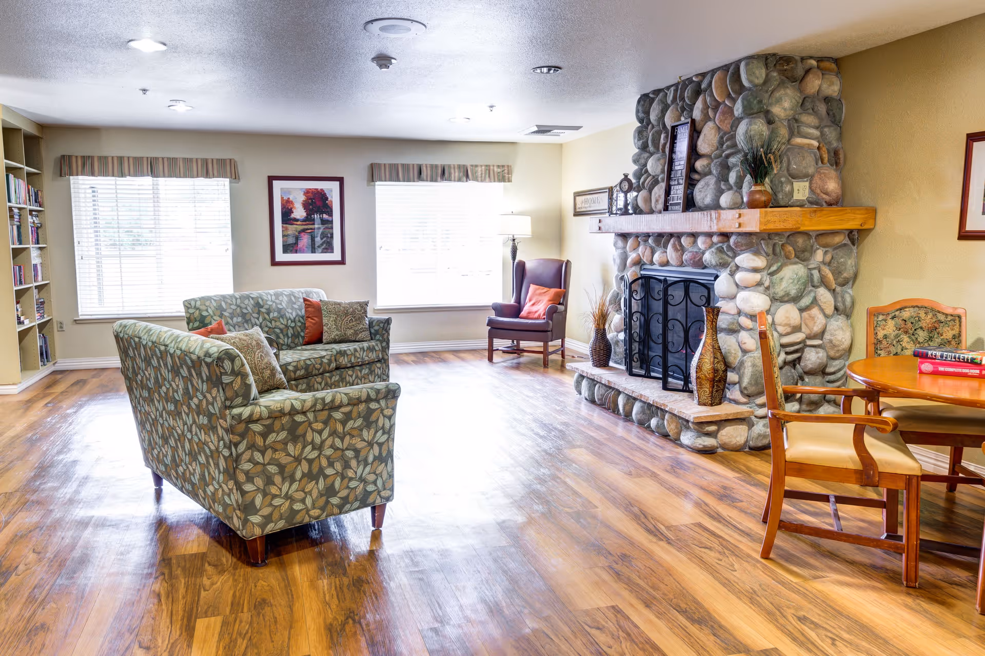 A cozy living room with wooden flooring, a stone fireplace with decorative vases on the hearth, a patterned sofa and loveseat with cushions, a wooden armchair with a red cushion, a round wooden table with chairs, bookshelves, and two large windows with blinds and valances letting in natural light.