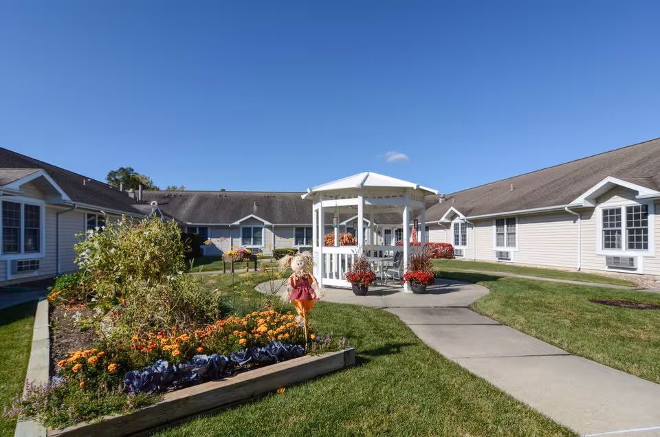 Outdoor courtyard area of a senior living community with a white gazebo in the center, surrounded by green grass, flower beds with orange and purple flowers, and a scarecrow decoration. The courtyard is enclosed by single-story beige buildings with multiple windows under a clear blue sky.