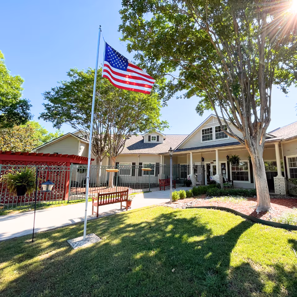 Front exterior of a senior living facility showing an American flag, benches, trees, and the building's covered entrance.