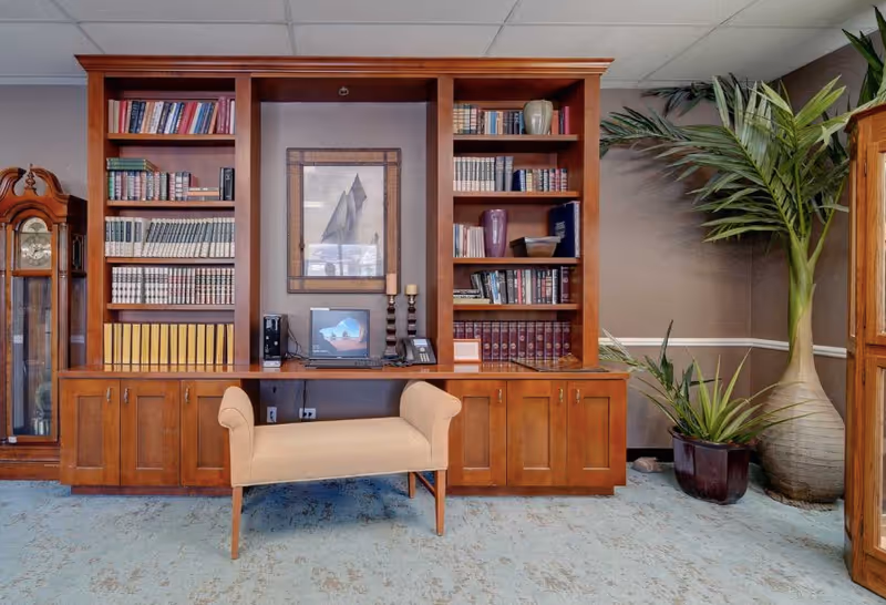 A cozy interior room featuring a wooden built-in bookshelf filled with books and decorative items. In front of the bookshelf is a beige upholstered bench. To the left, there is a tall grandfather clock, and to the right, two large potted plants. The room has a light blue carpet and neutral-colored walls with white trim.