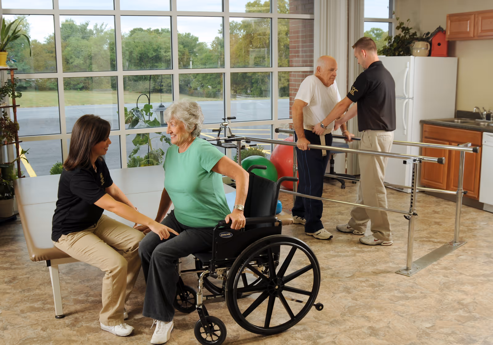 A senior woman in a wheelchair is assisted by a female caregiver in a bright room with large windows. In the background, a senior man is supported by a male caregiver while using parallel bars for physical therapy. The room has exercise equipment and a kitchenette area with a refrigerator and sink.
