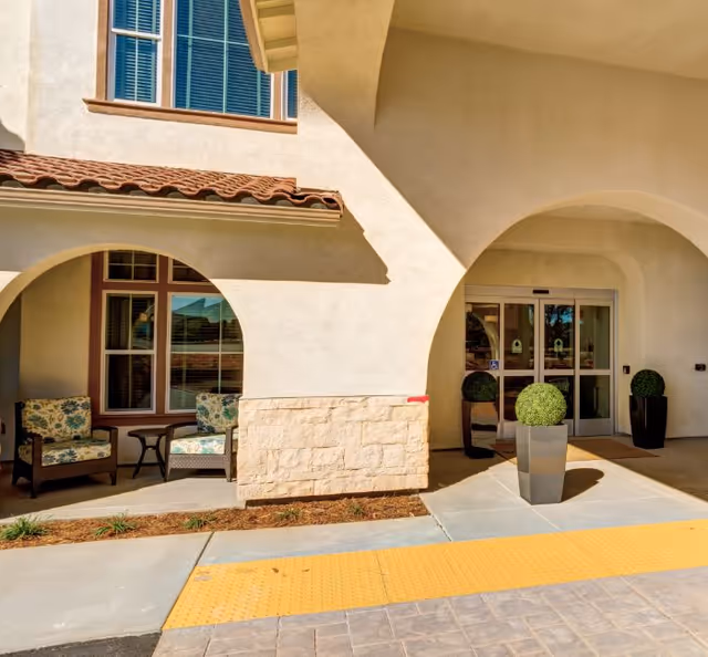 Front entrance of a light-colored building with arched covered entry, potted topiaries, and outdoor seating.