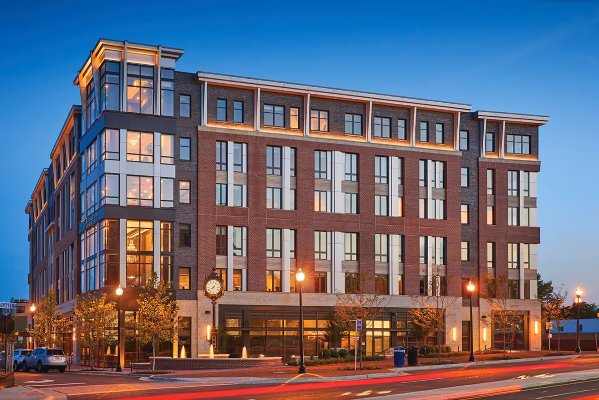 Exterior view of a modern multi-story senior living facility building named Cogir of West End Alexandria during dusk, with illuminated windows and street lamps, a clock on the sidewalk, and some trees and parked cars nearby.
