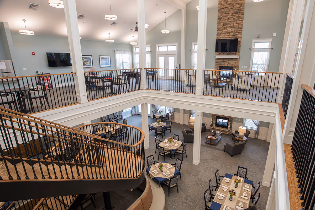 Interior view of a senior living facility with a two-story open space featuring a dining area with tables set for meals on the lower level and a lounge area with chairs and a fireplace on the upper level. The space has large windows with sheer curtains, light blue walls, and modern light fixtures. A staircase with black railings curves from the lower to the upper level.