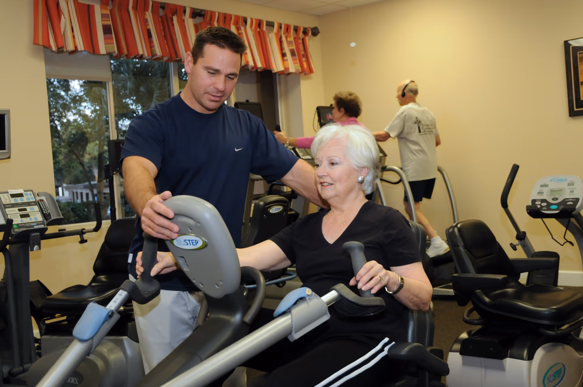 A man in a navy blue shirt assists an elderly woman with white hair who is using a NuStep exercise machine in a fitness room. In the background, two other elderly individuals are exercising on cardio machines near a window with red and white striped valances.