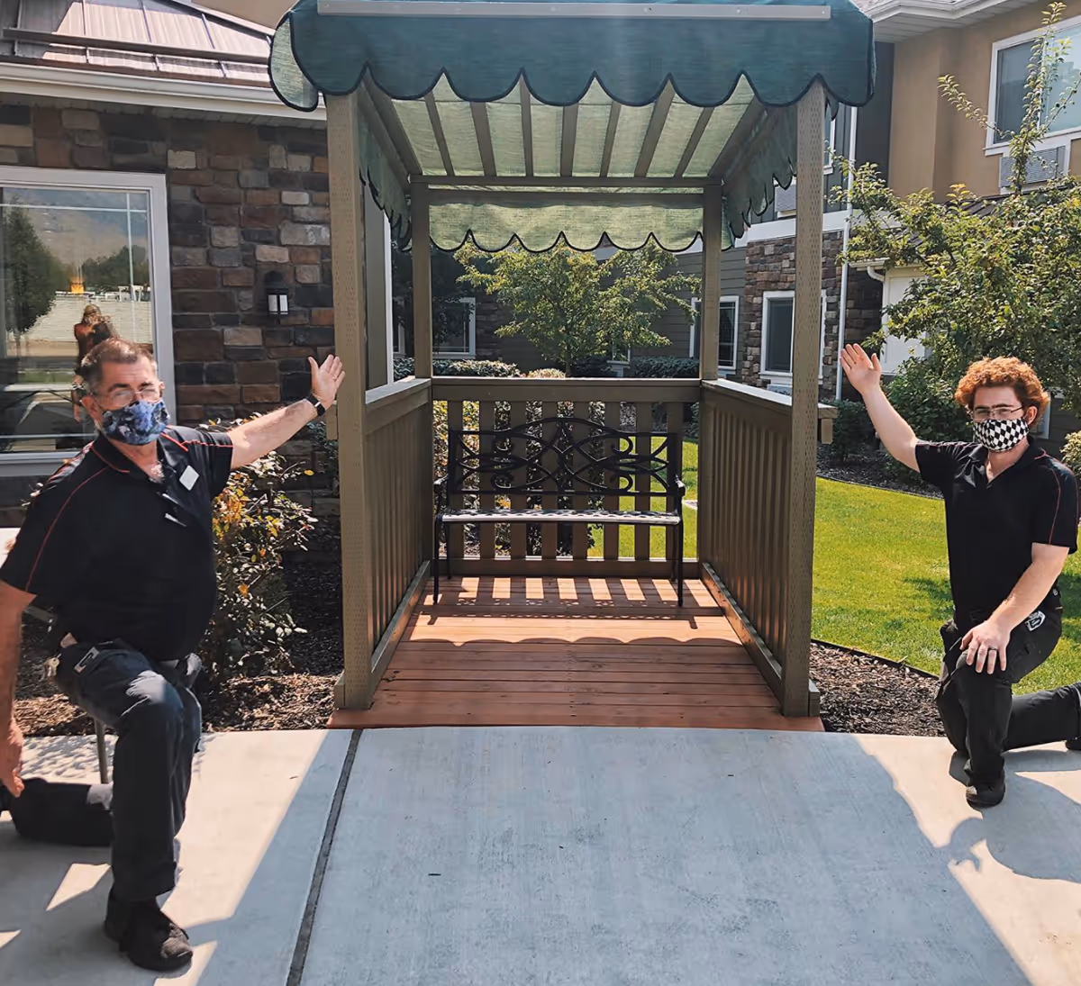 Two men wearing face masks kneel on either side of a small wooden gazebo with a green fabric roof and a decorative metal bench inside. They are outdoors on a concrete pathway with a building and greenery in the background.