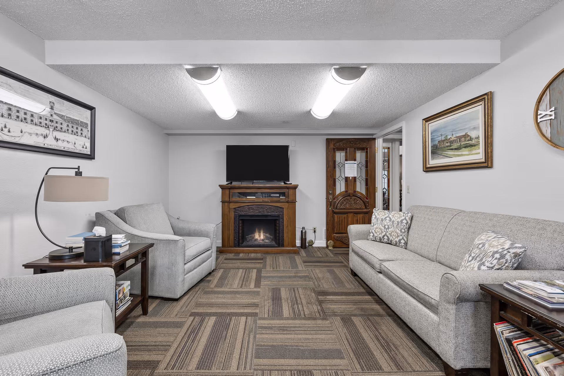 Cozy sitting room with gray sofas and armchairs facing a TV above an electric fireplace and a wooden door in the back.